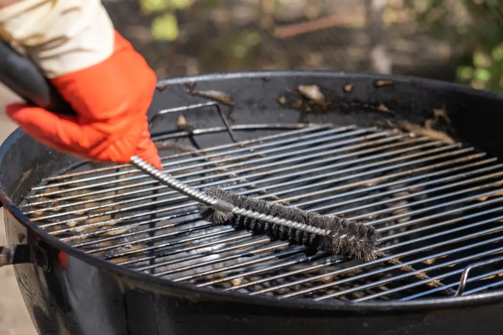 Man wearing orange gloves cleaning the grate of a charcoal kettle grill using a steel grill brush