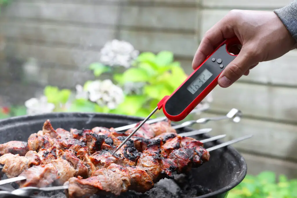 Man checking the temperature of beef kebabs with a digital meat thermometer on a charcoal grill
