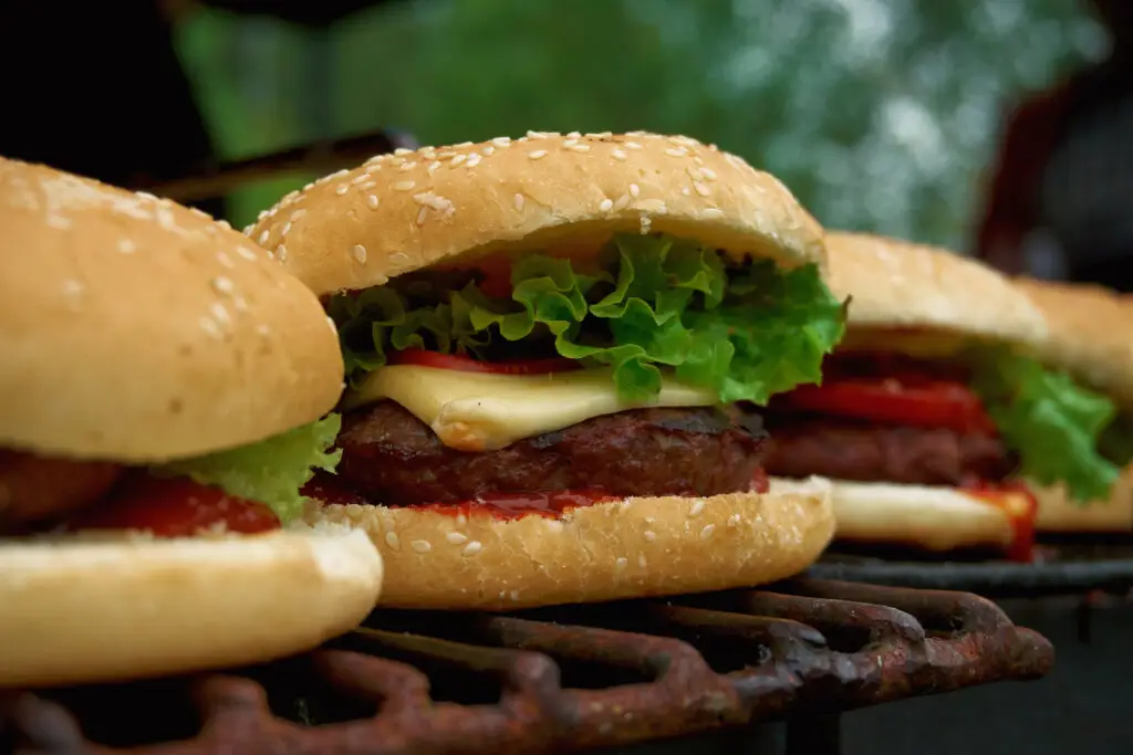 Grilled cheeseburgers with ketchup, lettuce, and tomatoes on sesame seed buns resting on a grill warming rack