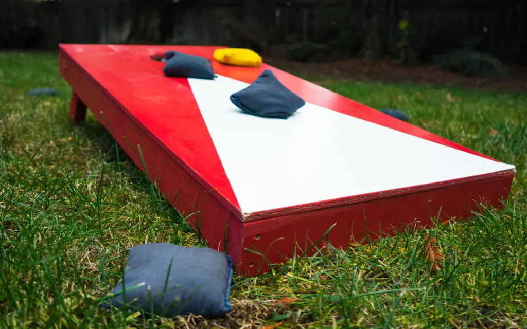 A red and white cornhole board with blue and yellow bean bags on the grass in a backyard