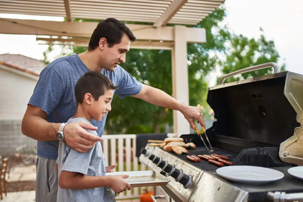 A father teaching his son how to grill hot dogs on a gas grill on the deck in the backyard