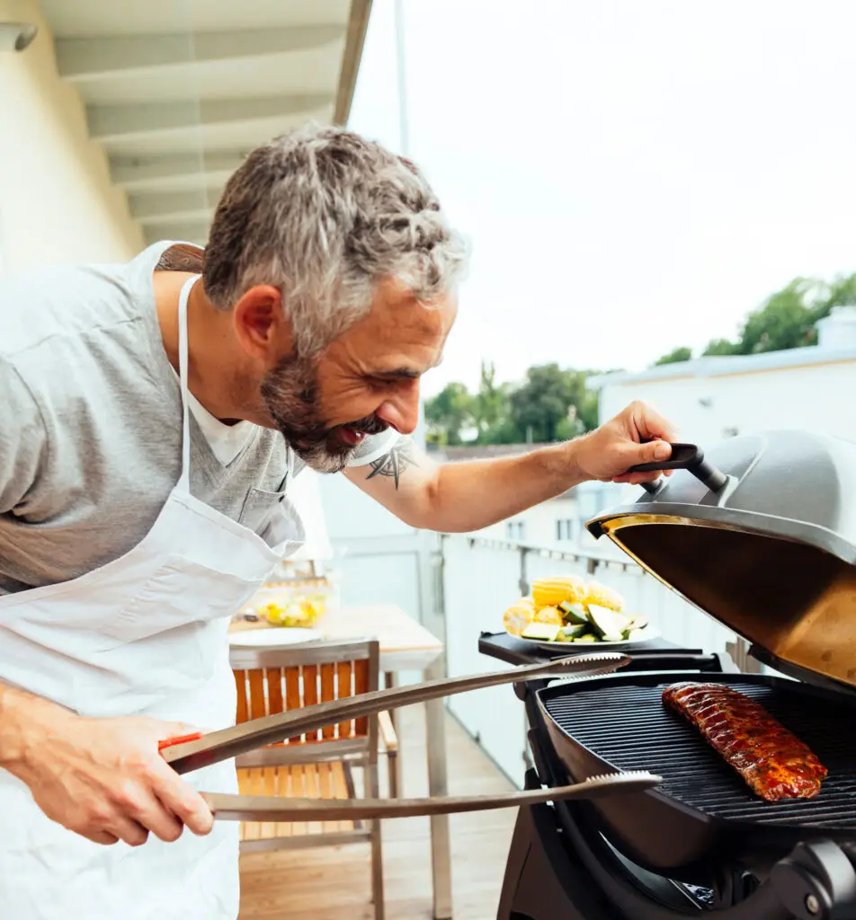 Man checking barbecue baby back ribs on a gas grill on a balcony