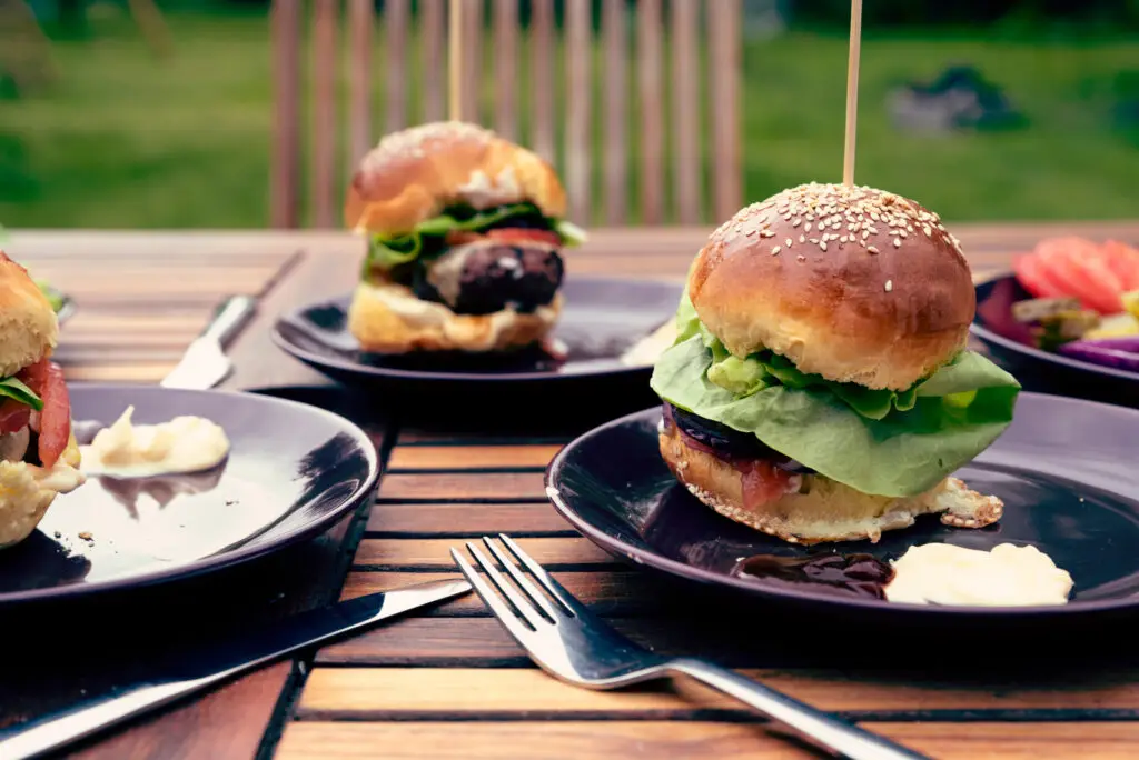 Bacon cheeseburgers with lettuce, tomato, pickles, and onions on sesame seed buns on a backyard picnic table
