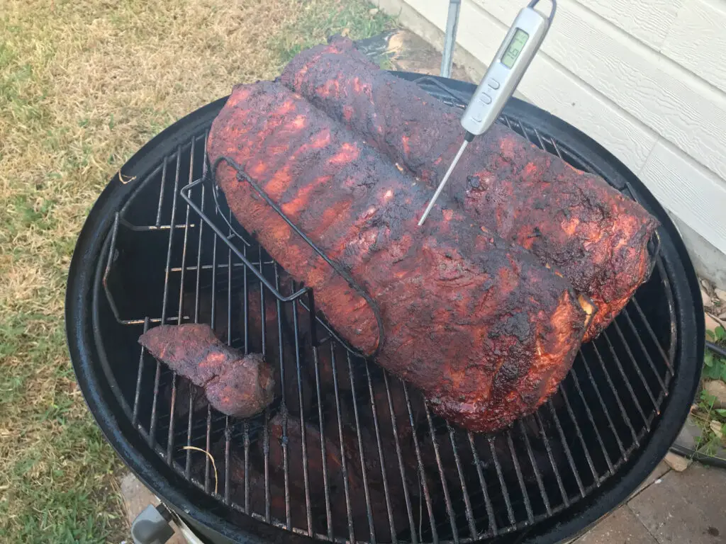 Pork ribs smoking on a charcoal smoker with a meat thermometer checking temperature